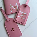 Three pink personalized luggage tags and a matching passport cover arranged on a light marble surface. The set features white monogram letters 'M' and 'C' with gold foil script and small gold airplane icons.