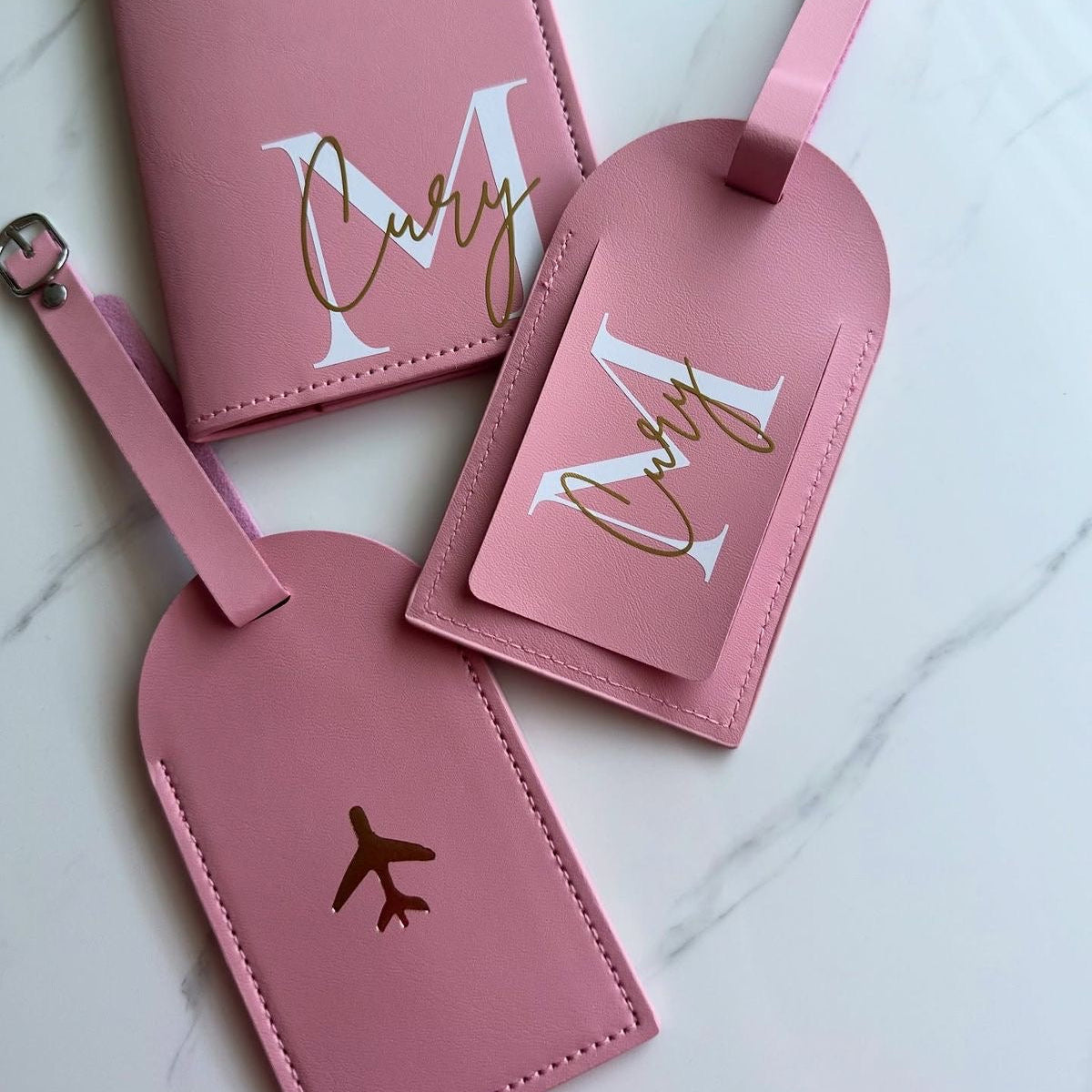 Three pink personalized luggage tags and a matching passport cover arranged on a light marble surface. The set features white monogram letters 'M' and 'C' with gold foil script and small gold airplane icons.