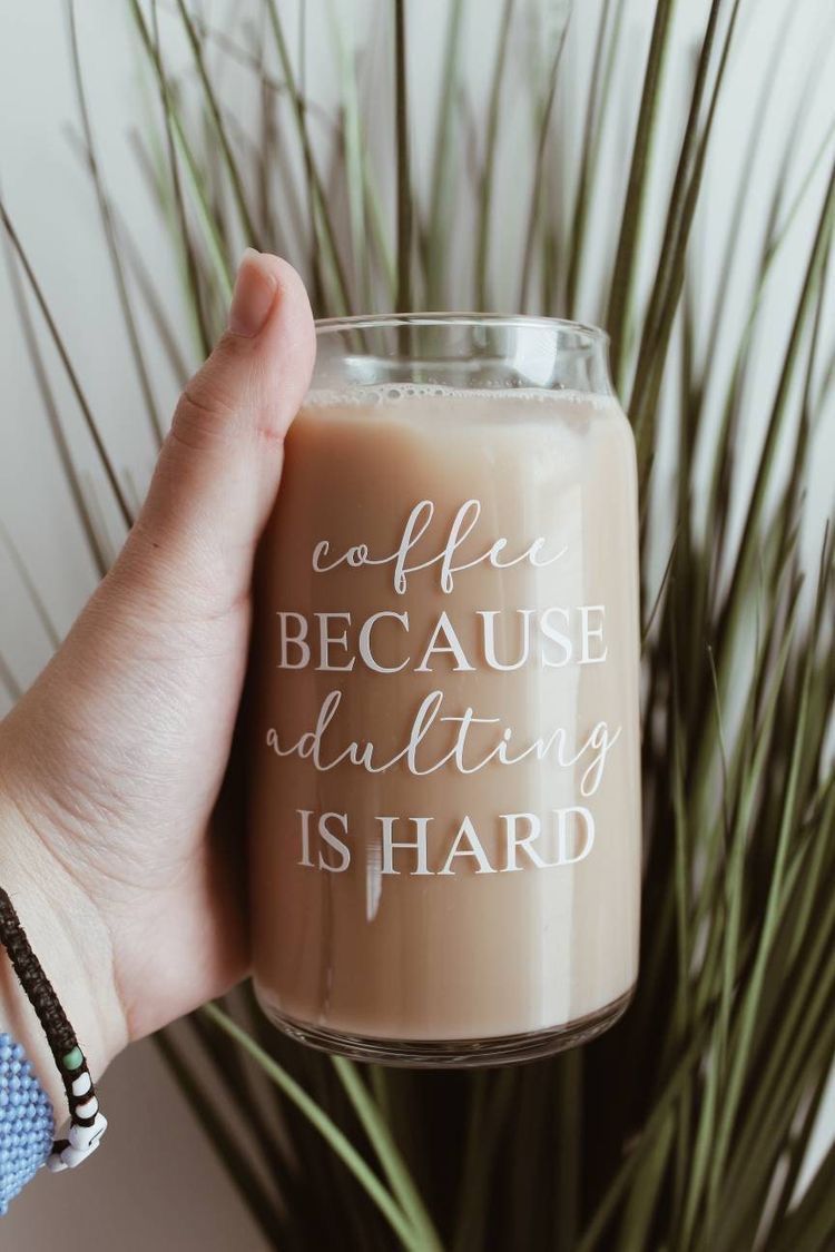 Glass of coffee with text 'coffee BECAUSE adulting IS HARD' held by a hand in front of greenery.