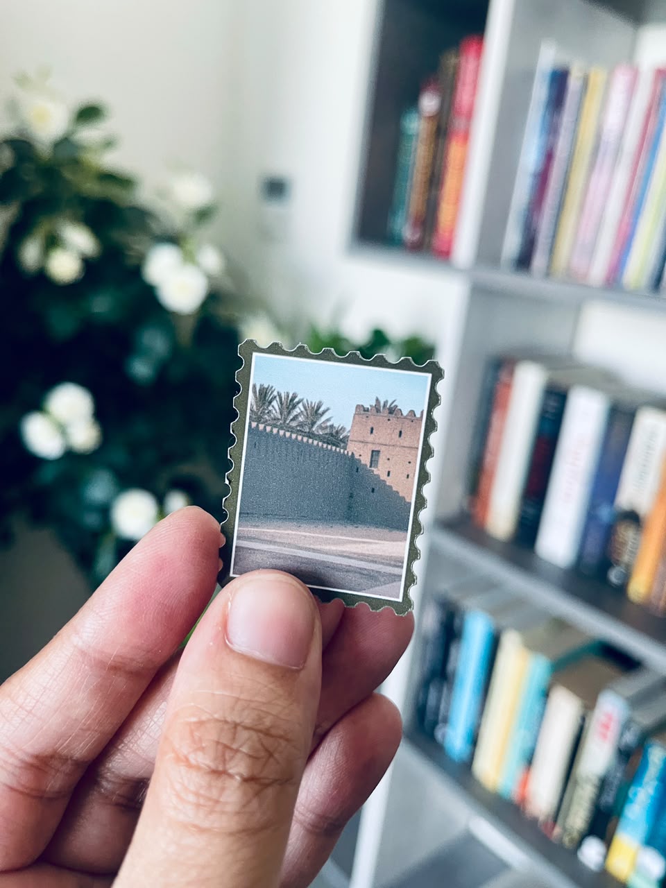A hand holding a small square postage stamp featuring a scenic photograph of a desert wall and tower, with blurred bookshelves and greenery in the background.
