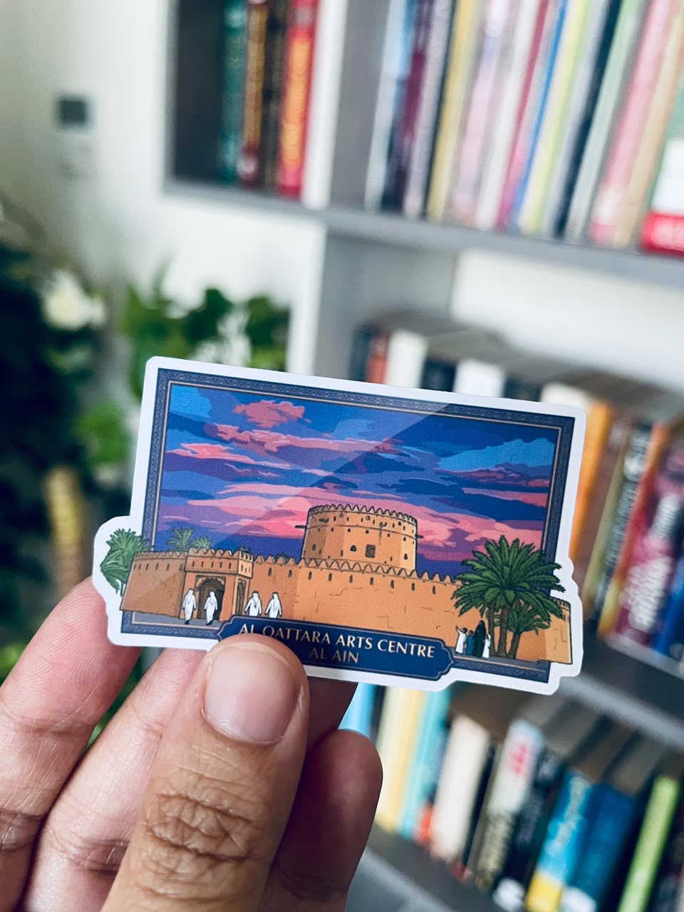 A hand holds a die-cut sticker featuring an illustrated view of Al Qattara Arts Centre in Al Ain, with a colorful sunset sky and a tower, against a blurred background of bookshelves.
