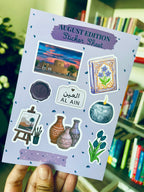 A hand holds a lavender sticker sheet titled 'AUGUST EDITION Sticker Sheet' featuring Al Ain themed illustrations, including a sunset scene, blue flowers, a seal stamp, a plaque reading 'AL AIN', pottery, a candle, and a small landscape painting, with a blurred bookshelf background.