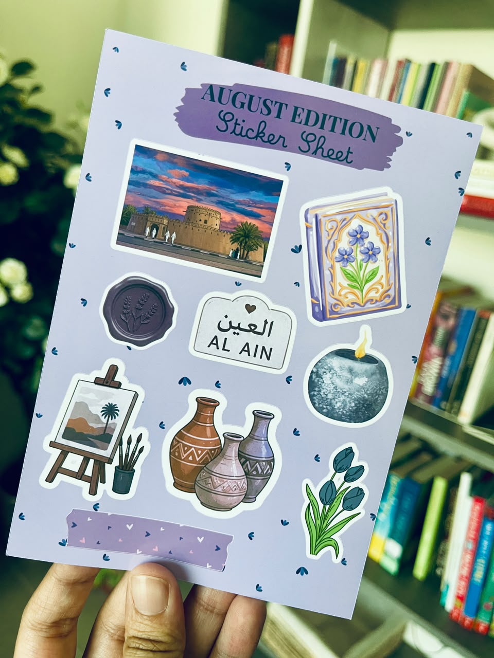 A hand holds a lavender sticker sheet titled 'AUGUST EDITION Sticker Sheet' featuring Al Ain themed illustrations, including a sunset scene, blue flowers, a seal stamp, a plaque reading 'AL AIN', pottery, a candle, and a small landscape painting, with a blurred bookshelf background.