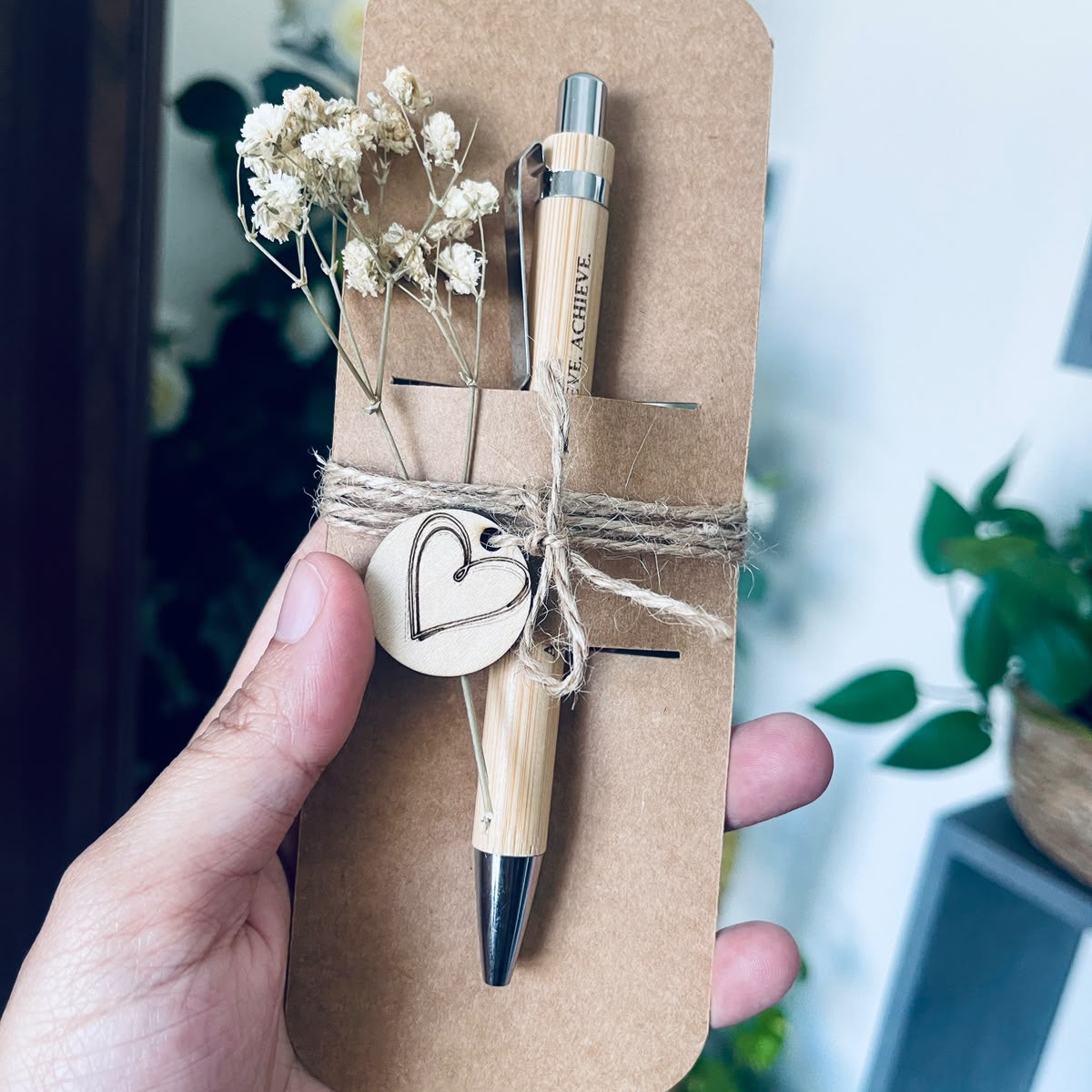 A hand holding a bamboo pen on a brown kraft paper card with dried flowers and twine, set against a softly blurred indoor background with green plants.