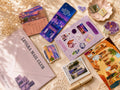 A flat-lay arrangement of stationery items on a beige fabric background with lace and dried flowers. Visible items include a lavender envelope printed 'LEVARA MAIL CLUB,' a purple calendar, a blue postcard, a lavender sticker sheet, small illustrated stickers, a postcard, a pen, and colorful illustrated cards or envelopes.
