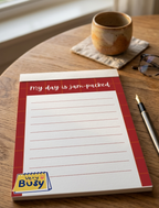 Notepad with 'My day is jam-packed' text on a wooden surface with a cup and glasses.